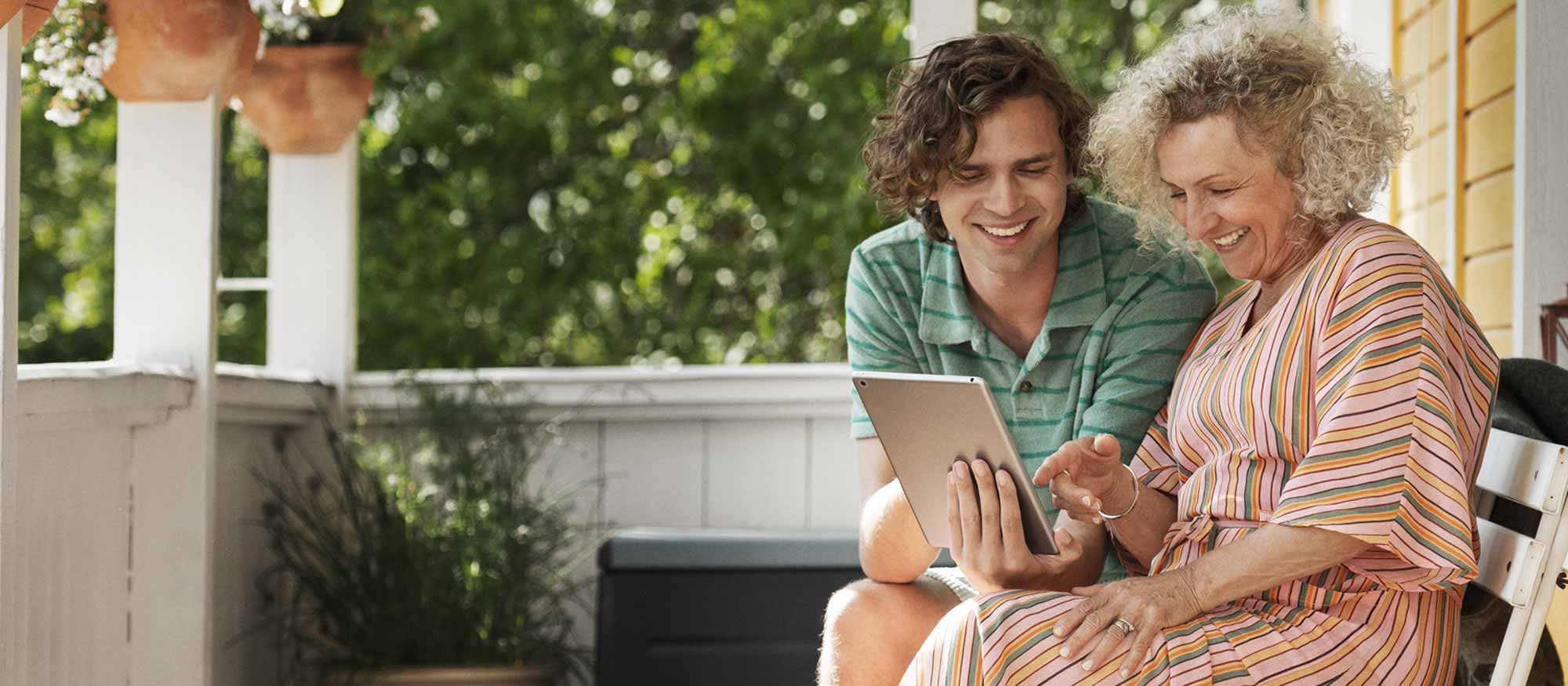 Yngre man visar äldre kvinna något på en surfplatta på verandan